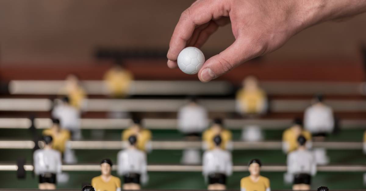 A close-up of a hand holding a white foosball above a foosball table. The foosmen are painted in yellow and white uniforms.