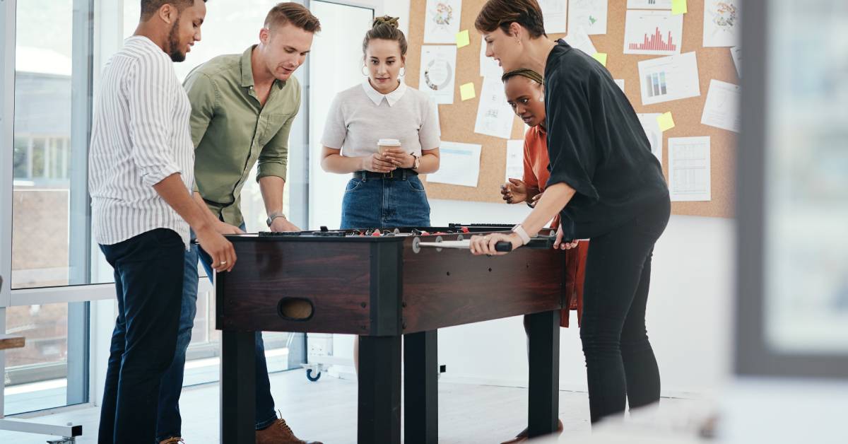 A group of office workers playing a game of foosball in front of a corkboard pinned with office paperwork and metrics.