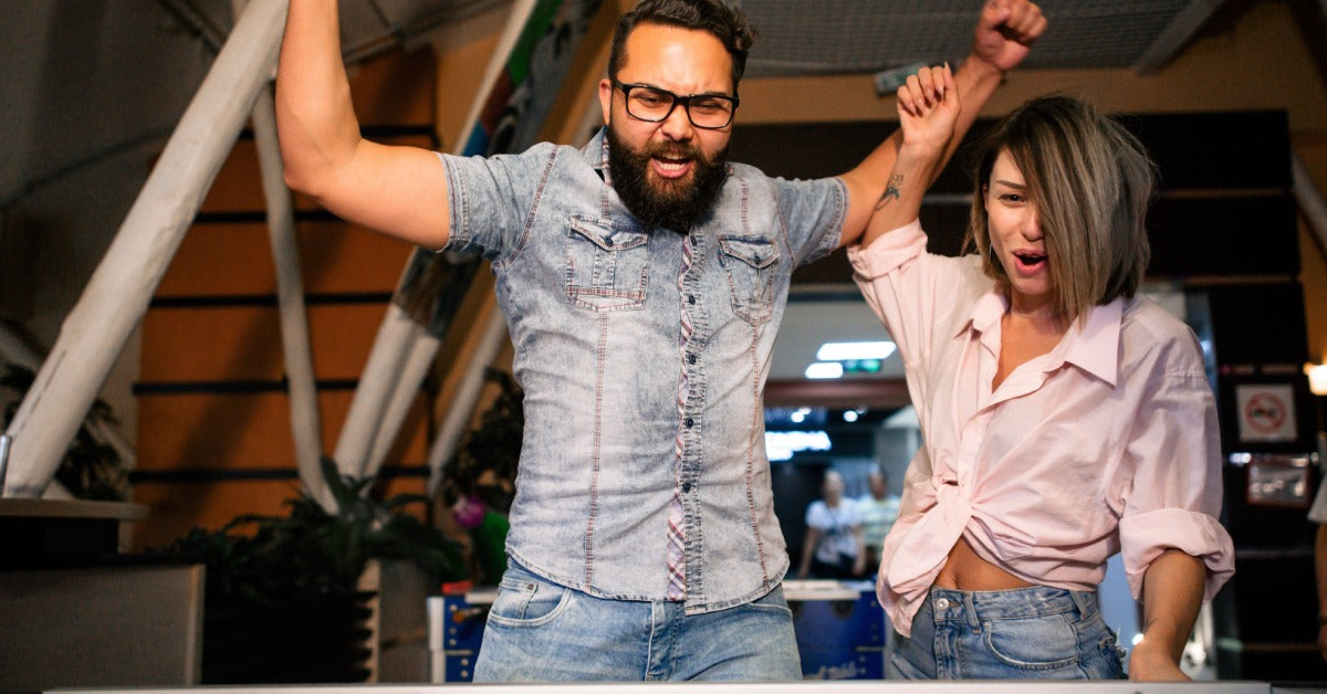 A man pumping his fists in the air and a woman cheering beside him. The edge of a foosball table is in the foreground.