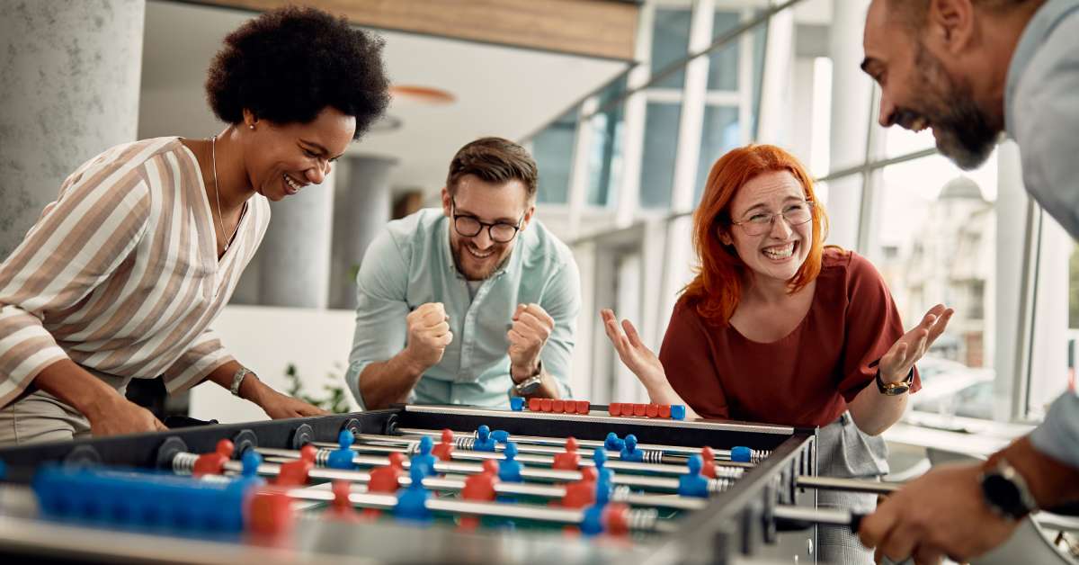 At a foosball table, two players excitedly compete against each other. Two friends cheer them on during the game.