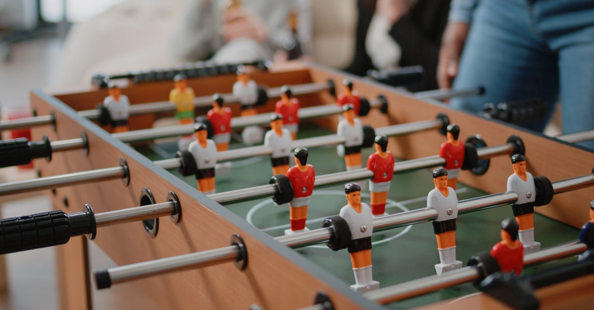 Close-up of a foosball table with red and white figurines. People stand behind the table and are blurred in the background.