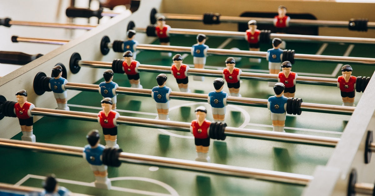 Foosball table from the side of the blue team versus the red team. Light reflects onto the green playing field.